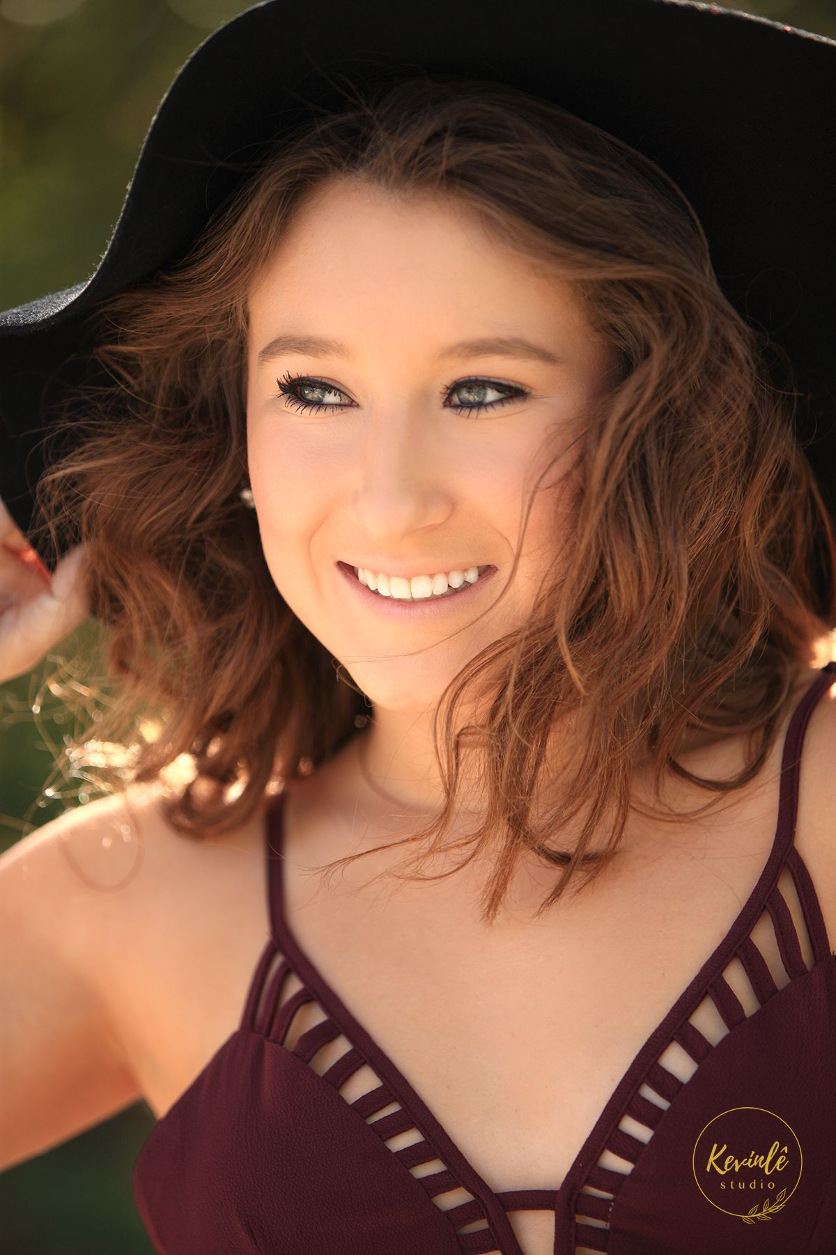 Smiling young woman with curly hair wearing a wide-brim black hat and burgundy top in a professional portrait.