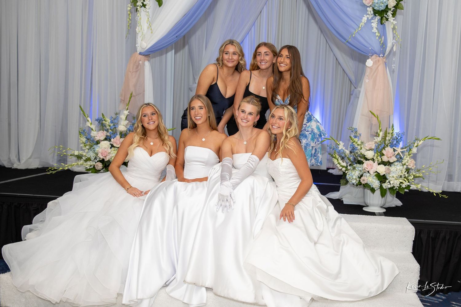 Six young women in elegant white ball gowns pose together at a formal debutante event with floral decorations.