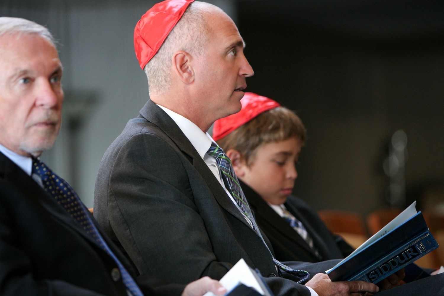 Three men wearing red kippot sit together during a Jewish religious ceremony, with one holding a prayer book.