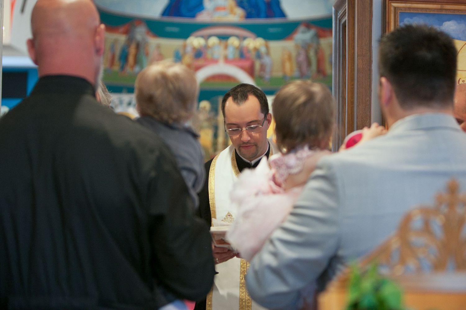 Priest in golden vestments performs a religious ceremony with attendees in a decorated Orthodox church.