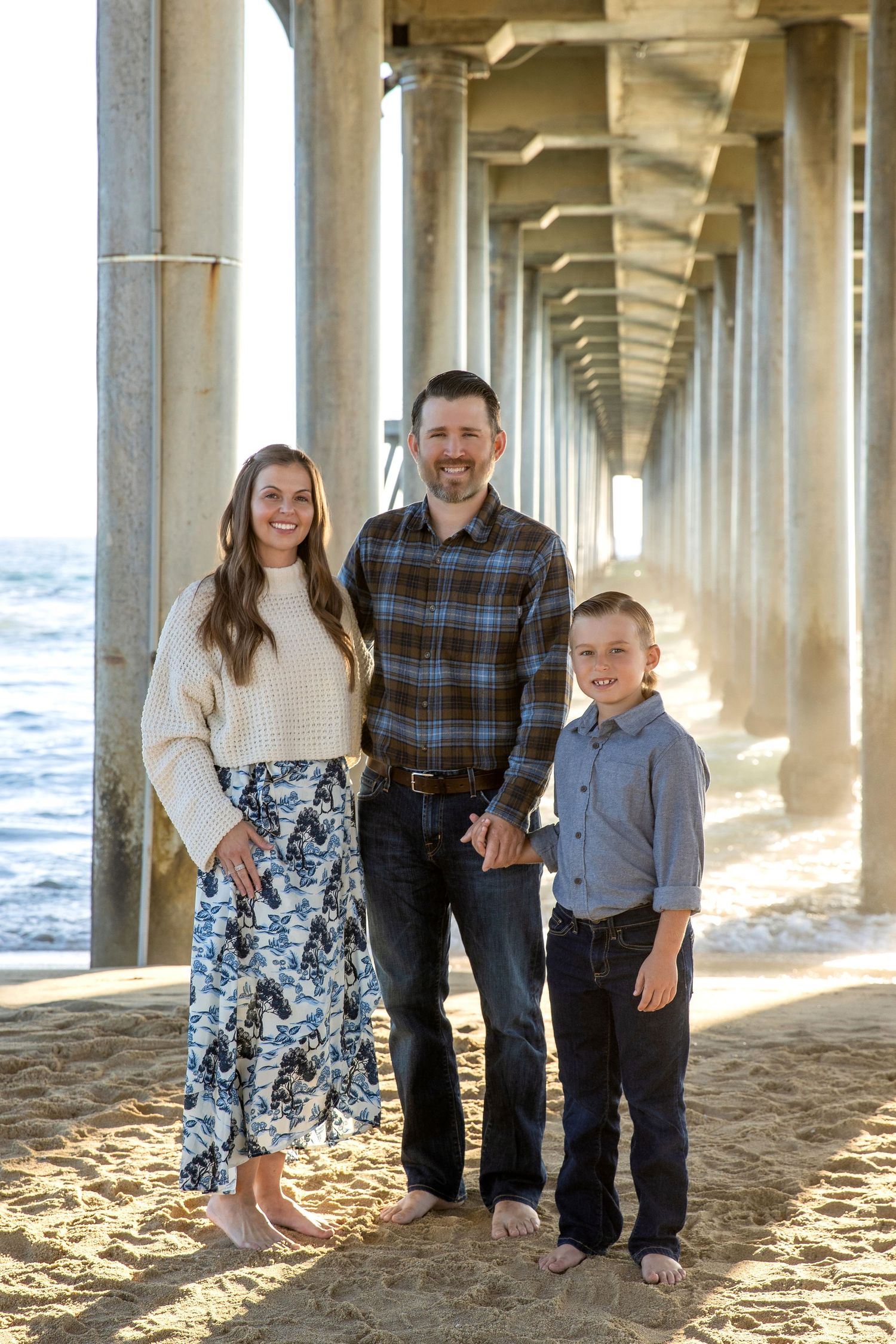 Family of three smiling under a beach pier on sandy shore, woman in floral skirt, man in plaid shirt, boy in blue.