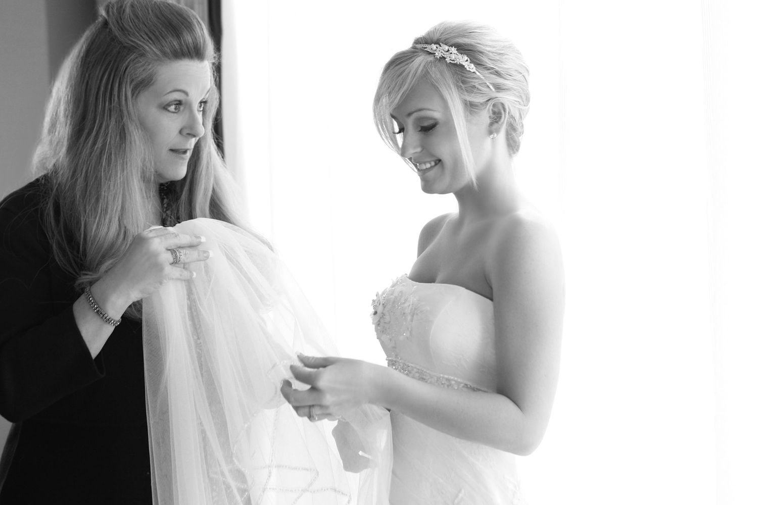 Black and white photo of a bride in a strapless gown having her veil adjusted by a smiling bridesmaid.