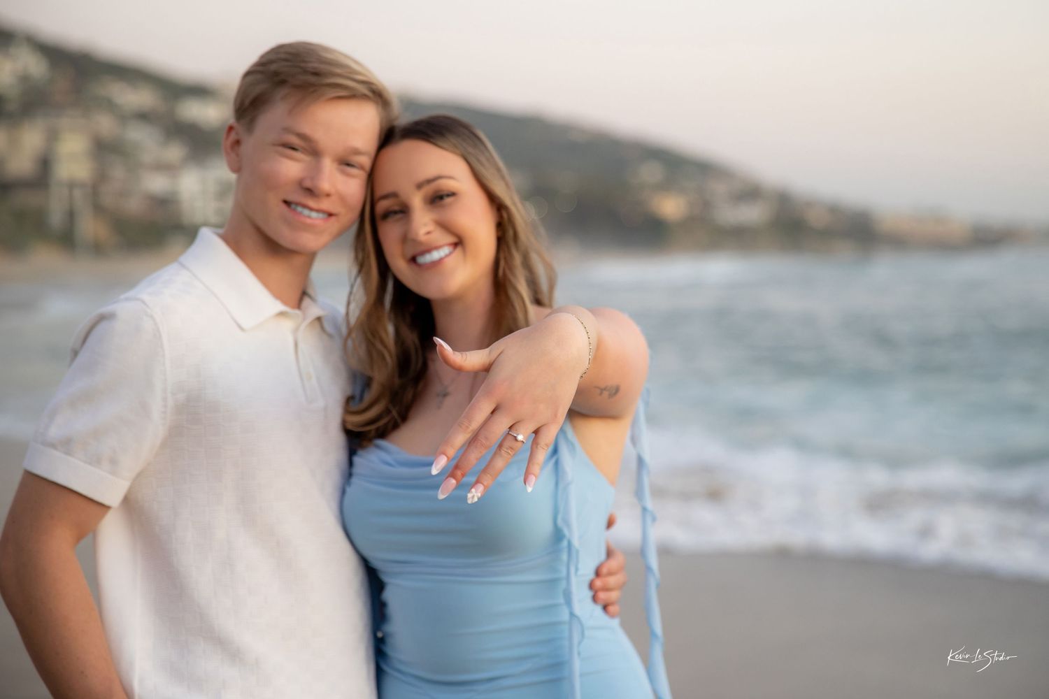 Happy engaged couple posing on beach, woman showing off engagement ring.