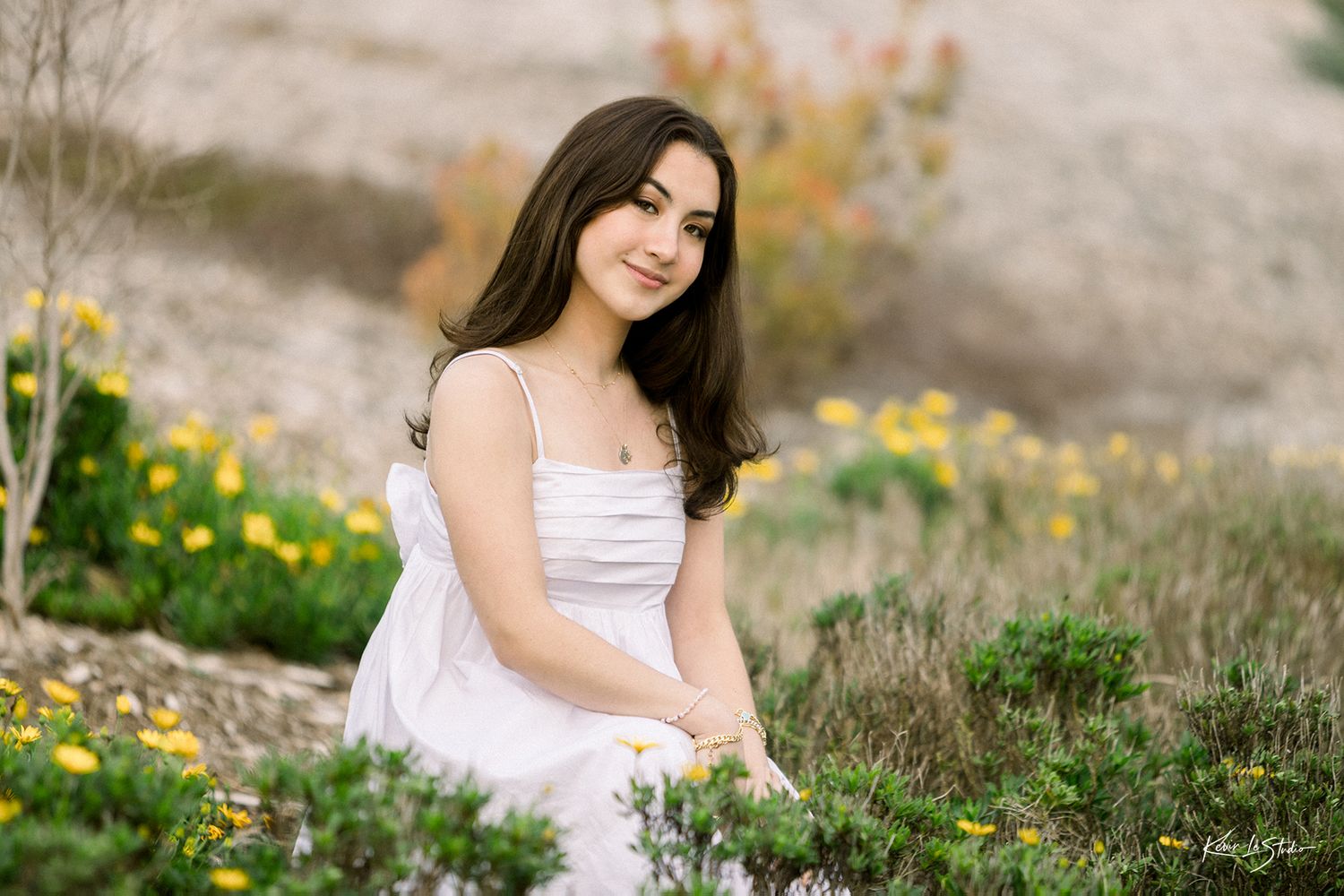 Young woman in white dress sits smiling among yellow flowers in a natural outdoor setting.