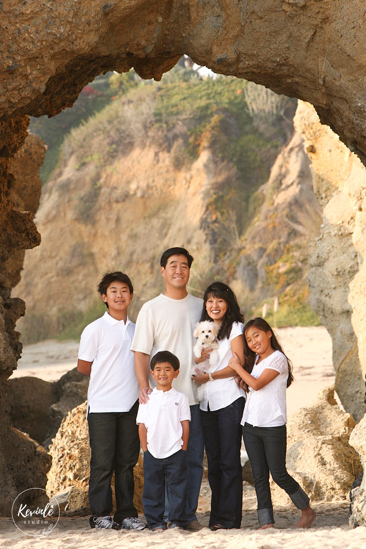 Family of five posing under a natural rock arch on a sandy beach, wearing matching white tops.