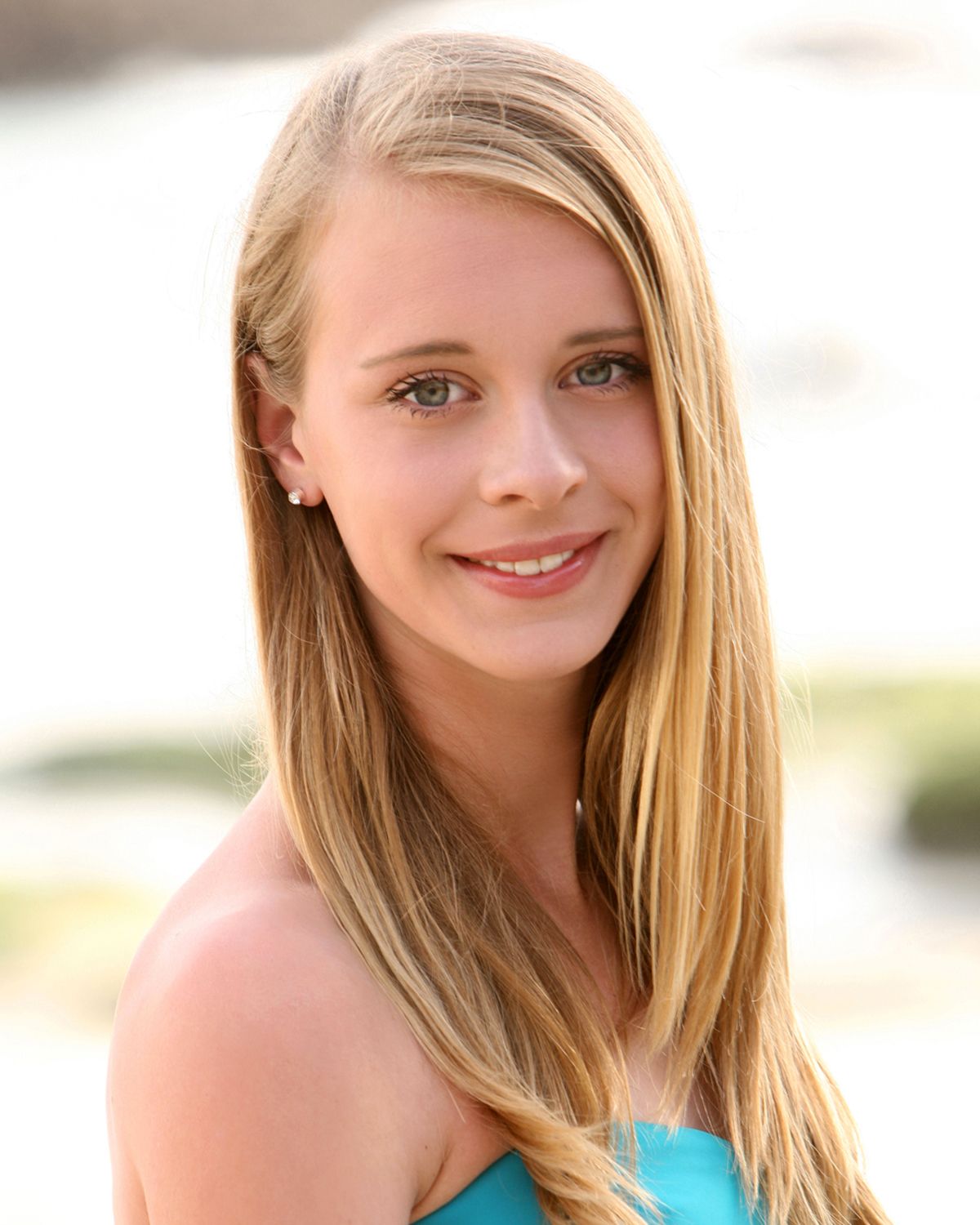 Smiling young blonde woman in turquoise top posing outdoors with blurred background.