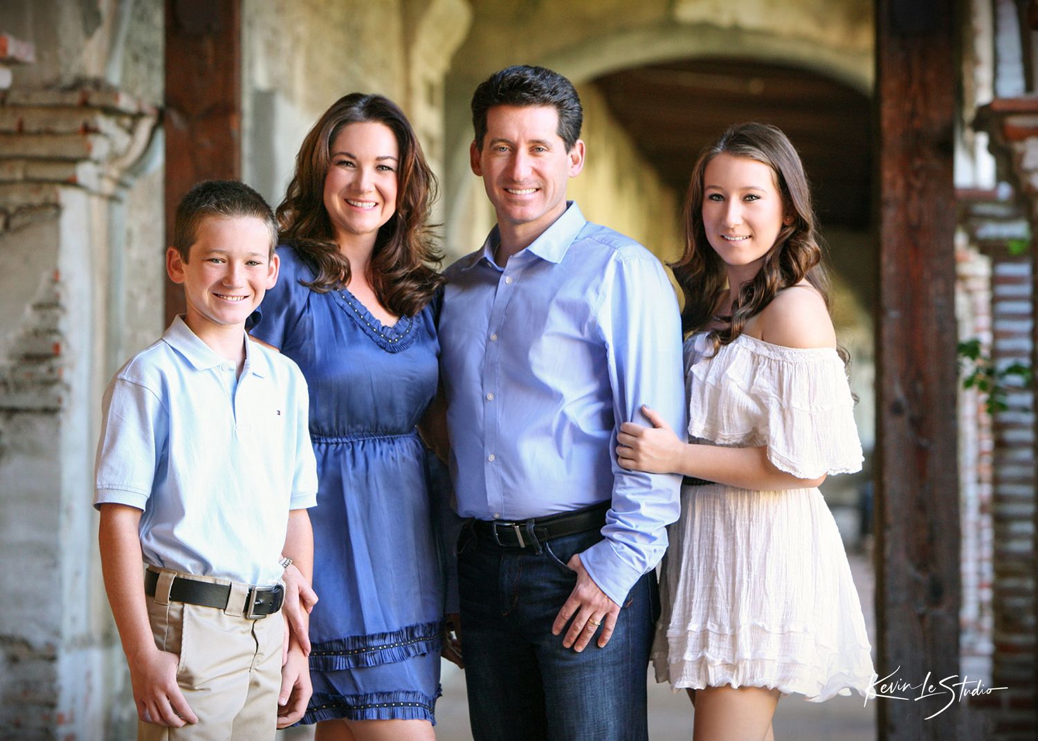 A smiling family of four poses together in a rustic archway setting, wearing casual elegant attire.