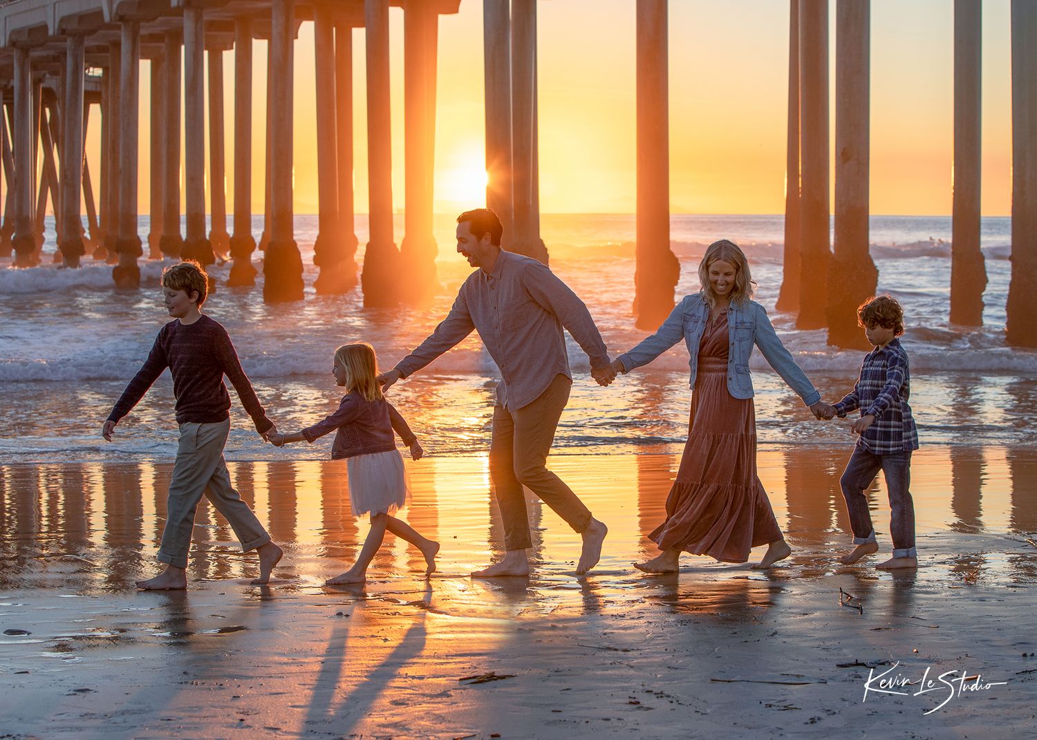 Family of five holding hands walking on beach at golden sunset near pier, waves reflecting warm orange light.