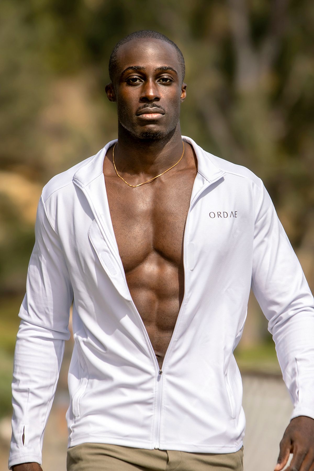 Muscular Black man wearing open white ORDAE zip jacket with gold chain necklace outdoors in natural setting.