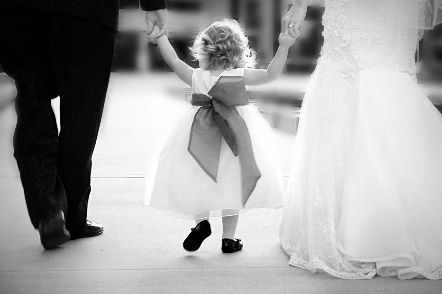 Black and white photo of a flower girl in a white dress with bow, holding hands with bride and groom at a wedding.