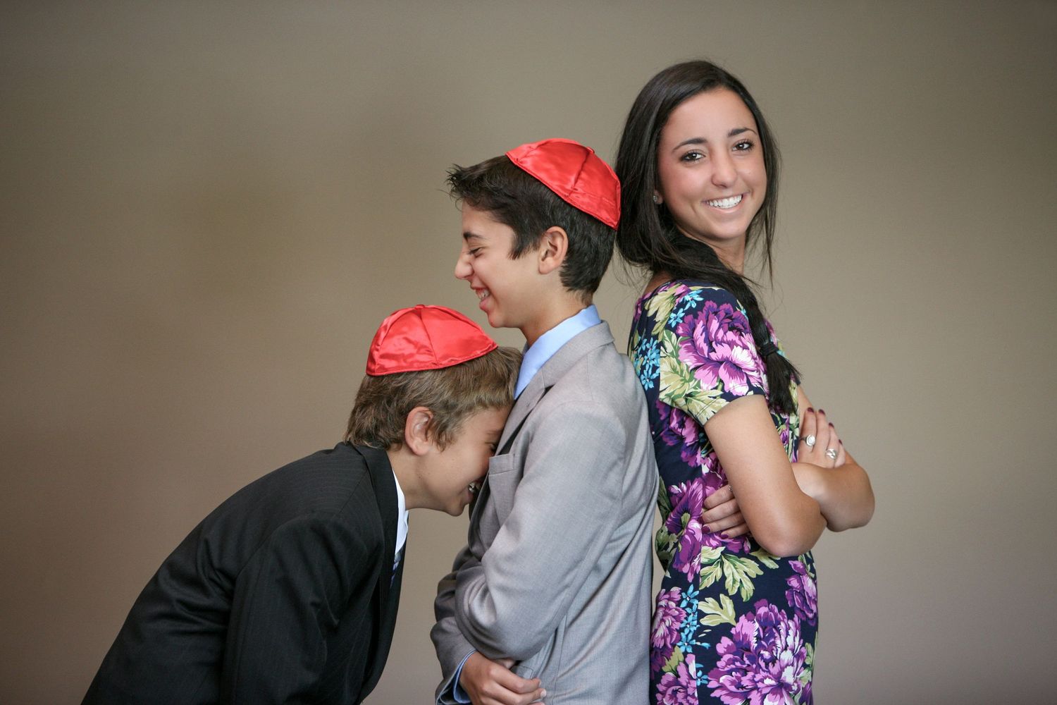 Two boys wearing red kippot pose with a smiling young woman in a floral dress for a formal portrait.