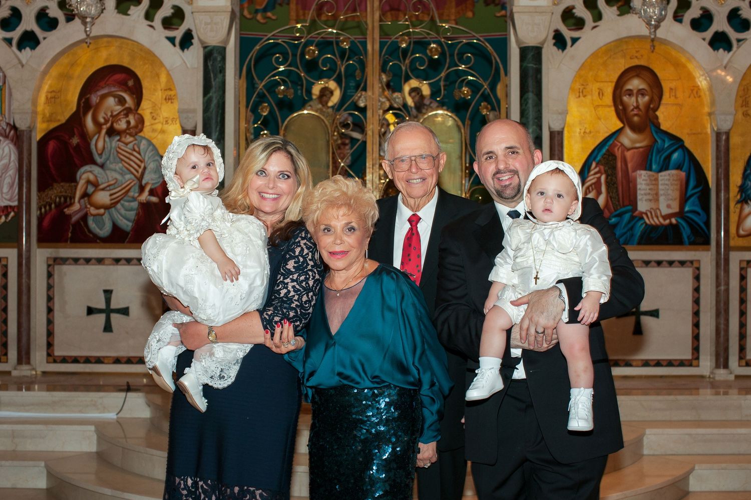 Family portrait in ornate church with two adults holding babies in white christening gowns, flanked by older couple.