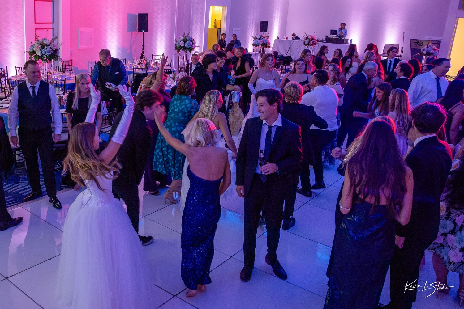 Guests dancing at an elegant wedding reception with pink and blue ambient lighting on a white dance floor.