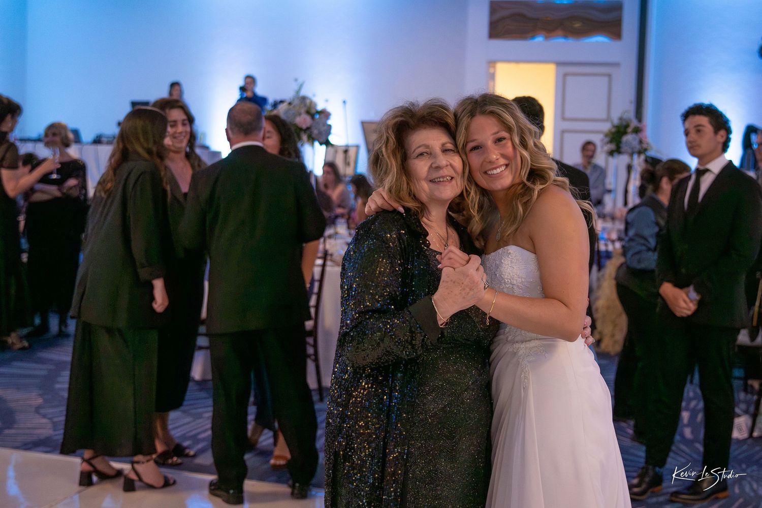 Two women smile and embrace at an elegant wedding reception, one in a white bridal gown and the other in a dark sequined dress.