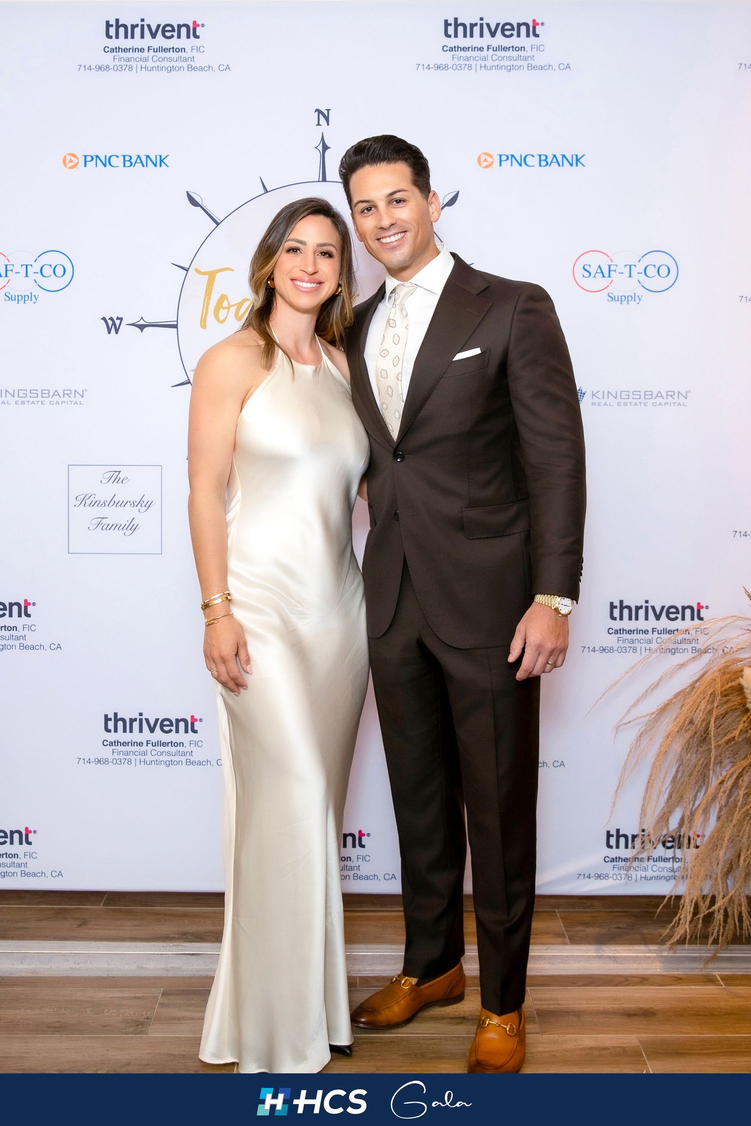 Couple posing at HCS Gala event, woman in white satin gown and man in brown suit, in front of sponsor backdrop.