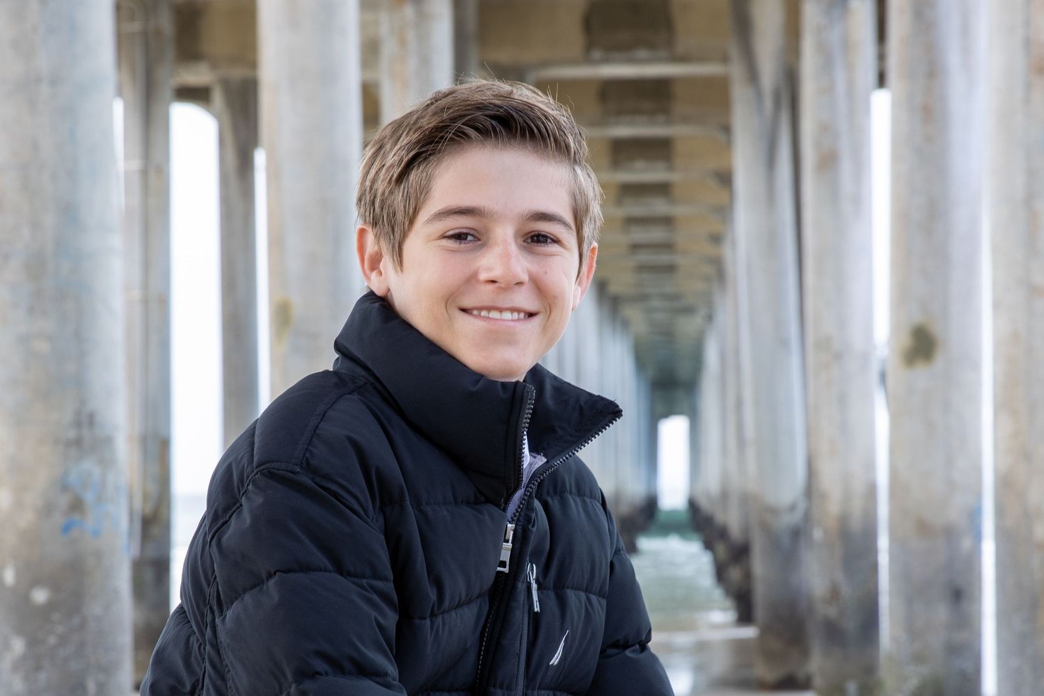 Smiling teenage boy in black jacket poses under a pier with columns stretching into the background.