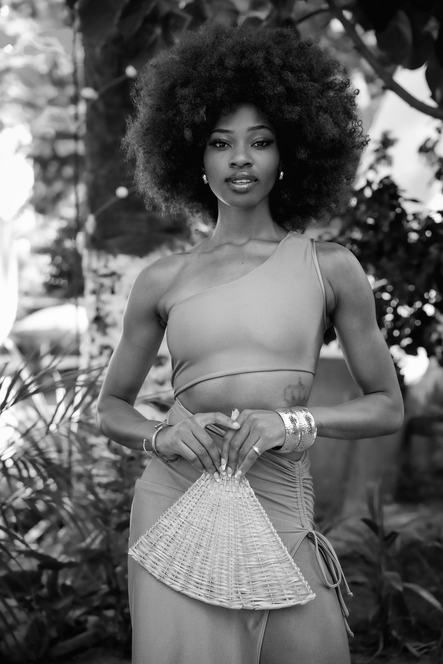 Black and white photo of woman in crop top holding wicker fan in garden.
