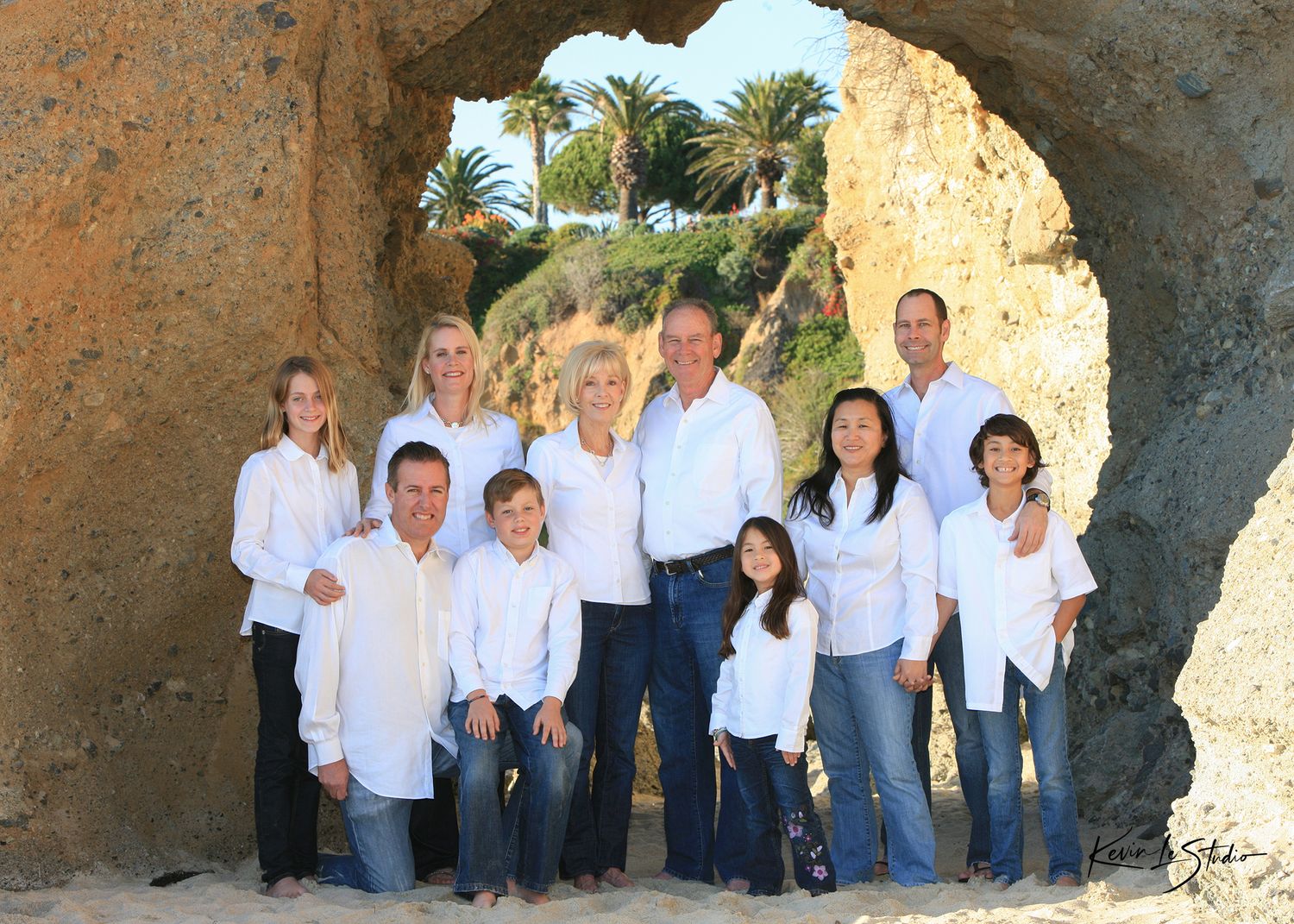 Large family group portrait posing under a natural rock arch on a sunny beach, all wearing white tops and jeans.