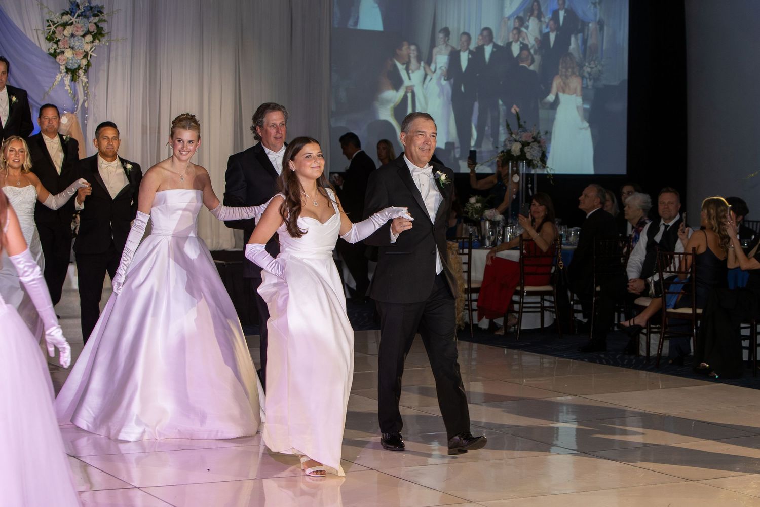Elegant debutante ball with young women in white gowns and long gloves dancing with escorts on a grand ballroom floor.