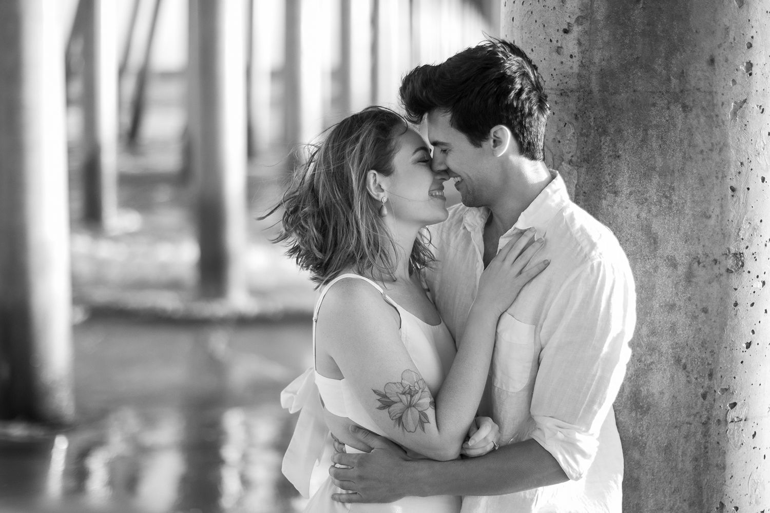 Black and white photo of a romantic couple embracing and about to kiss under a pier near the water.