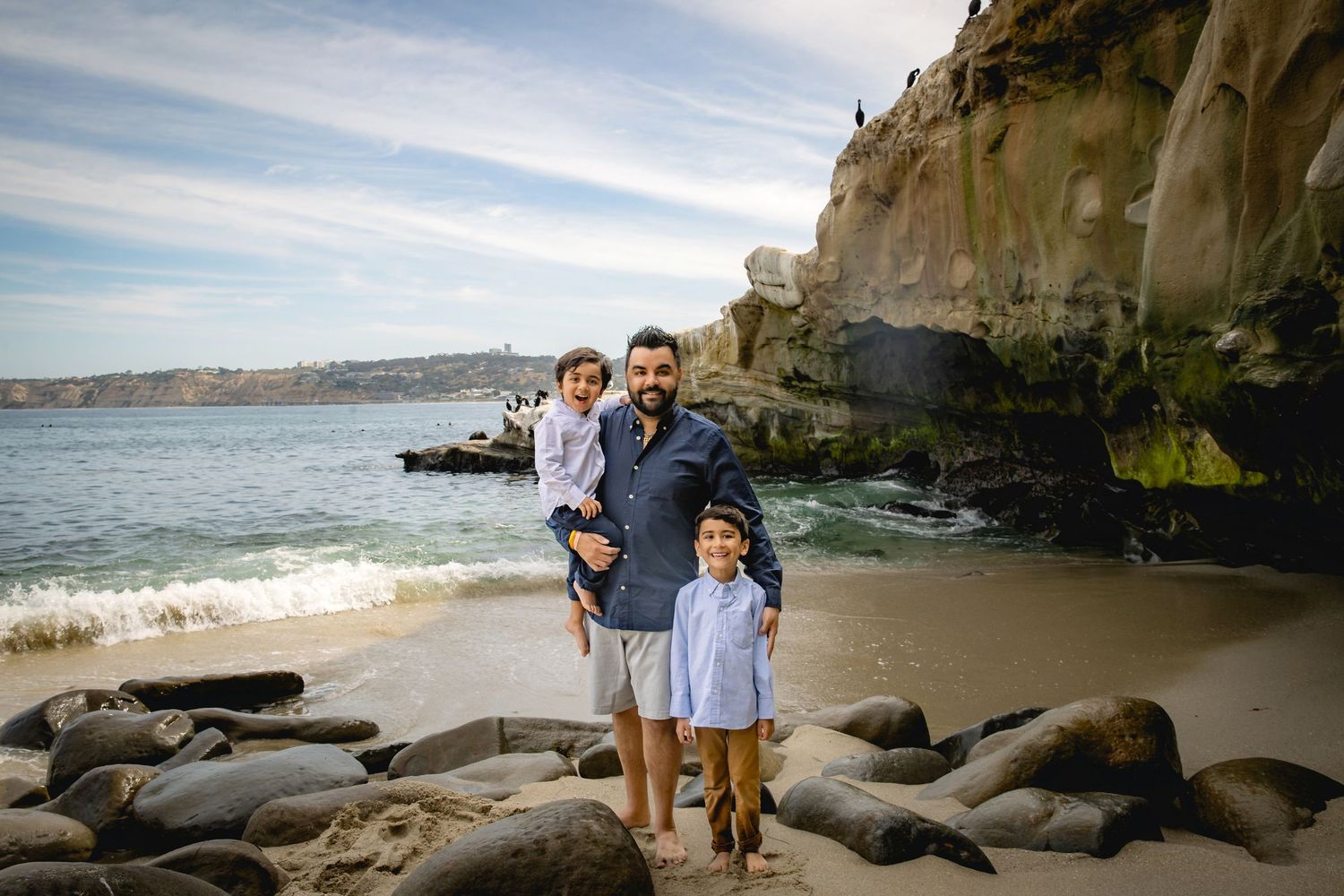 A father holds his young child while standing with another child on a sandy beach with rocky cliffs.