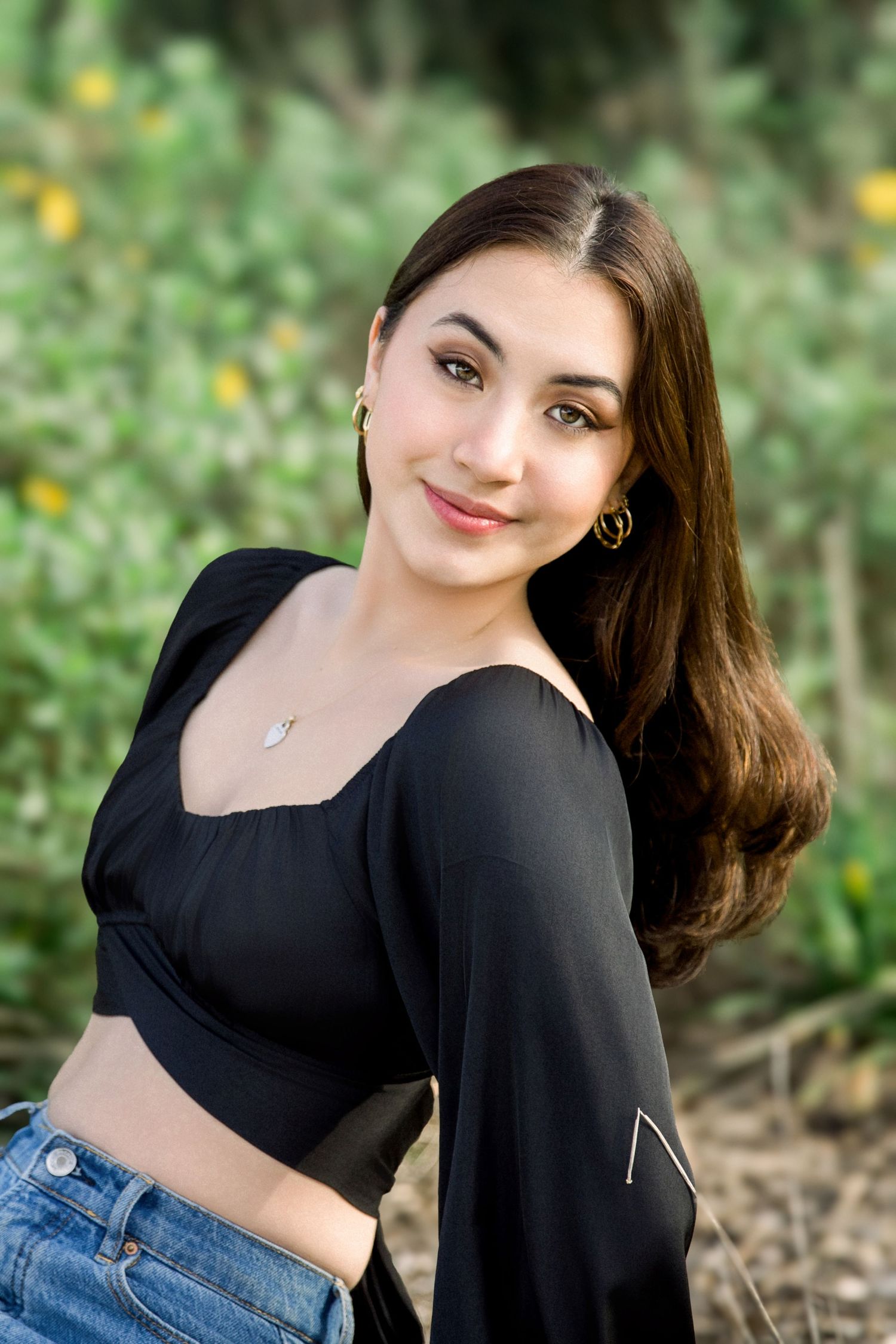 Young woman in black crop top and jeans smiles outdoors, wearing gold hoop earrings and a delicate necklace.