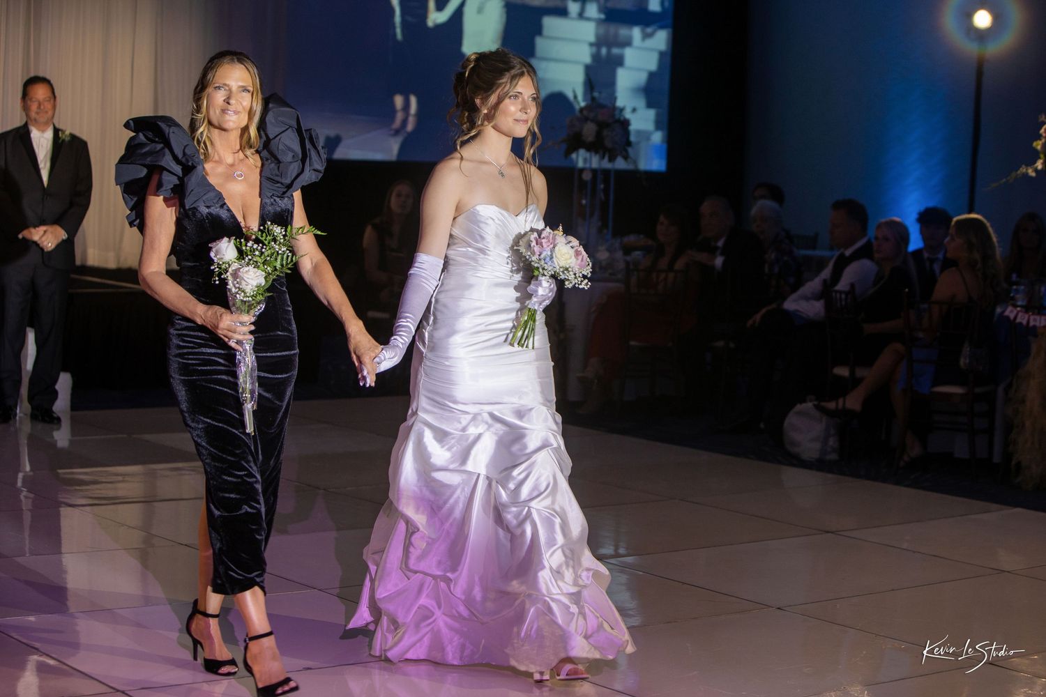 Two women walk a runway, one in a black gown and one in a white ombre wedding dress, both holding bouquets.