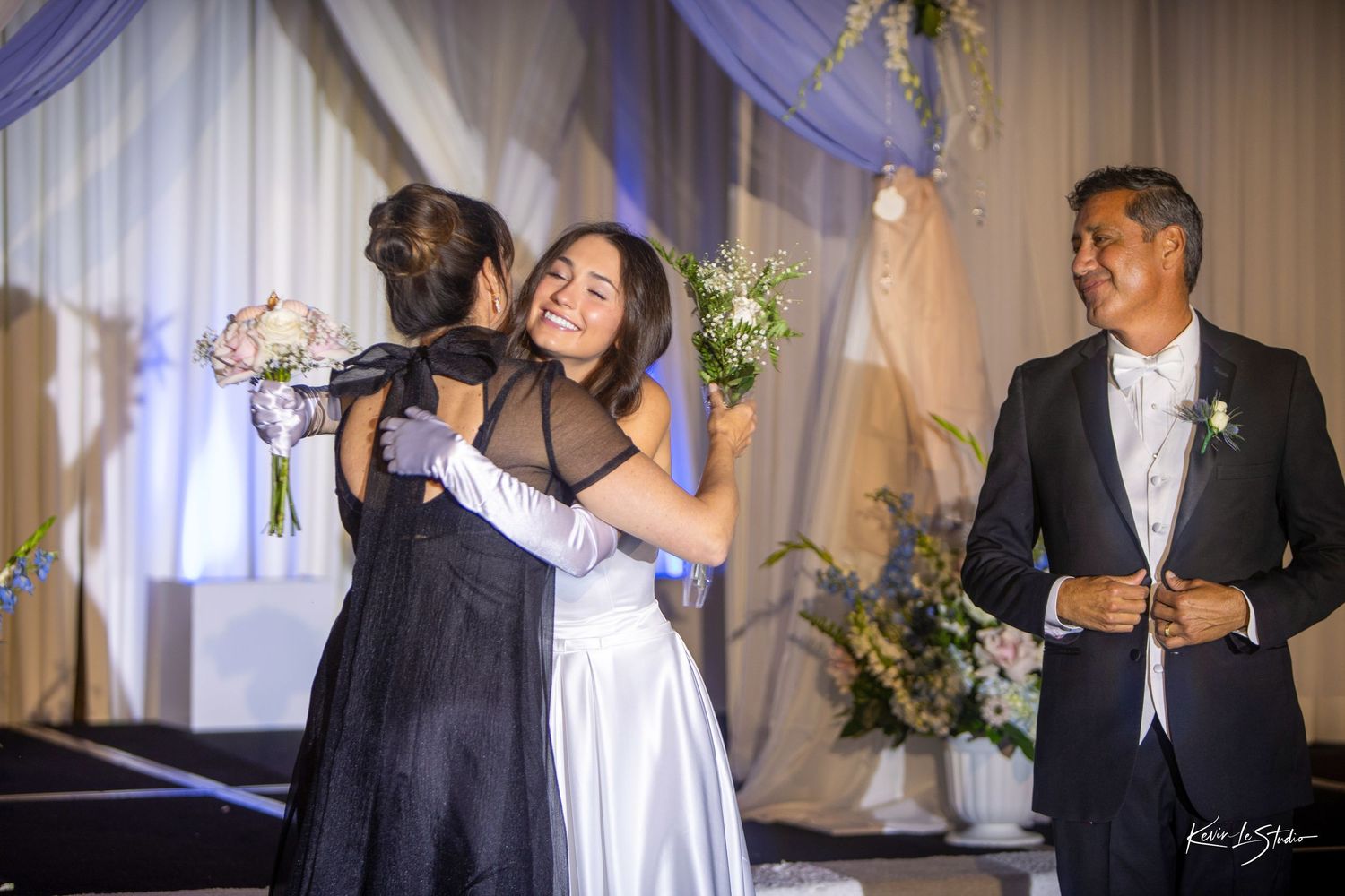 Bride in white gown hugs guest in black dress at wedding reception while father of the bride smiles nearby.