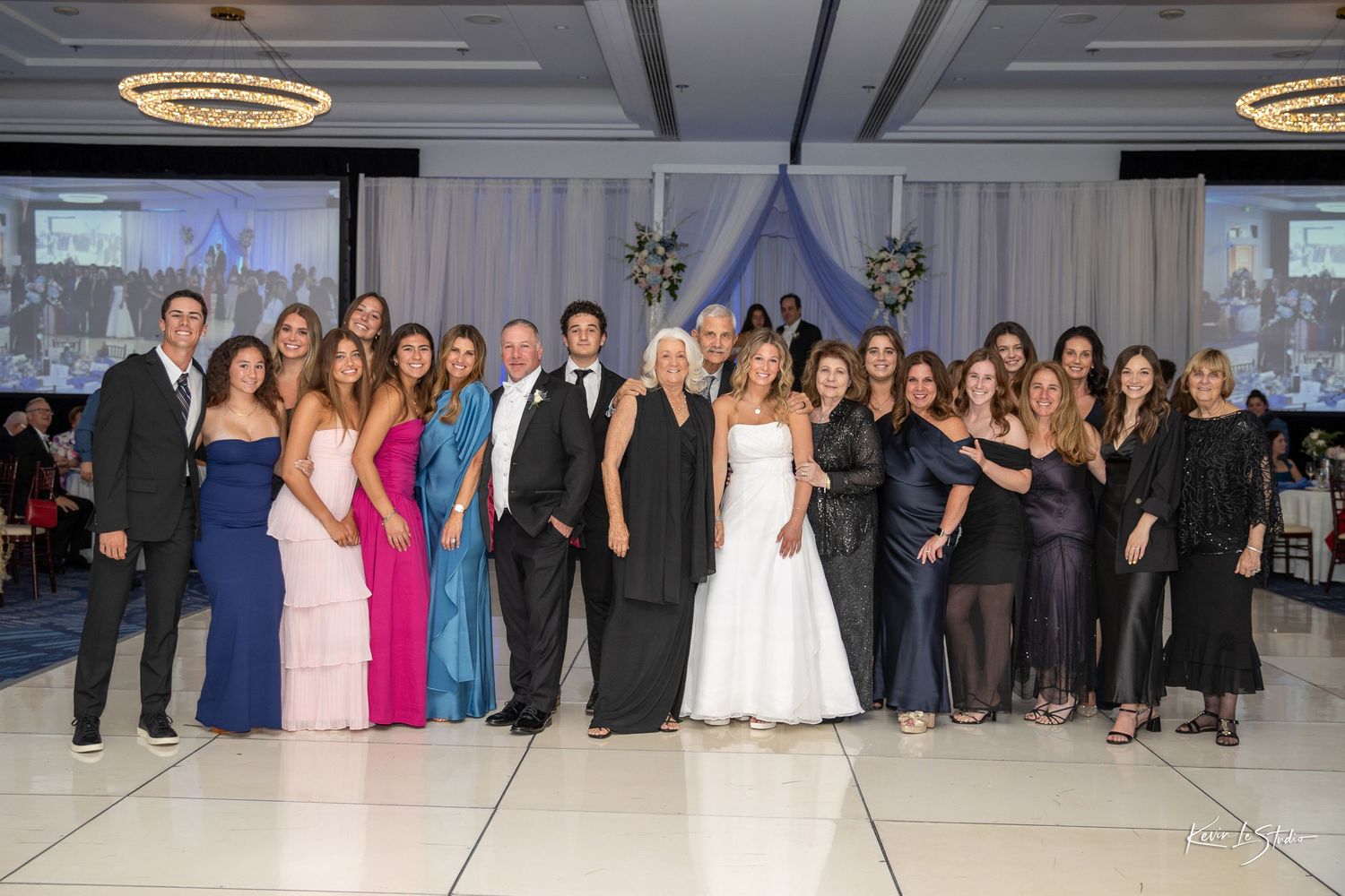 Wedding party poses together in elegant ballroom with blue draping, bride in white gown surrounded by guests in formal attire.