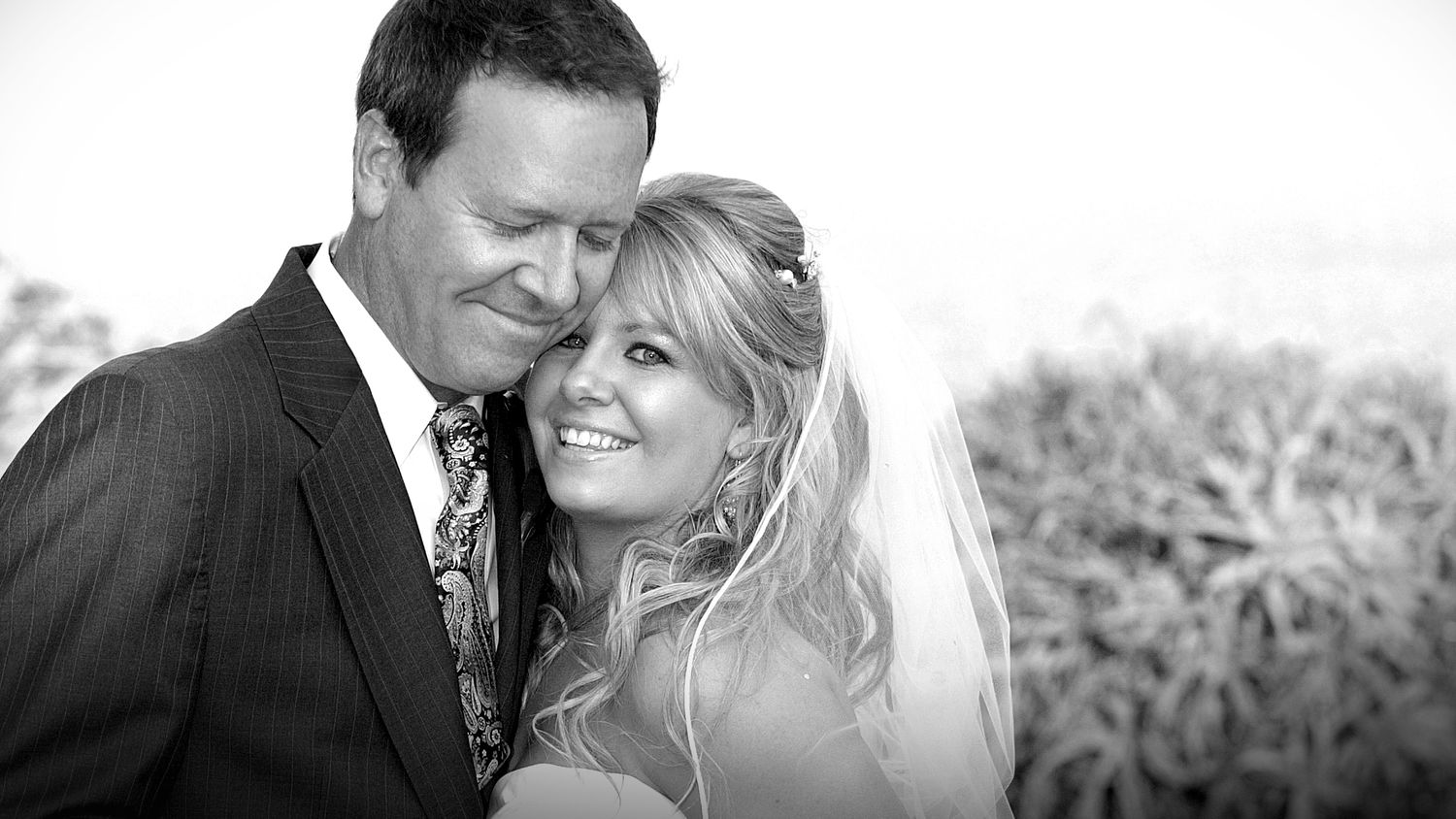 Black and white wedding photo of smiling bride in veil and groom in suit embracing outdoors.