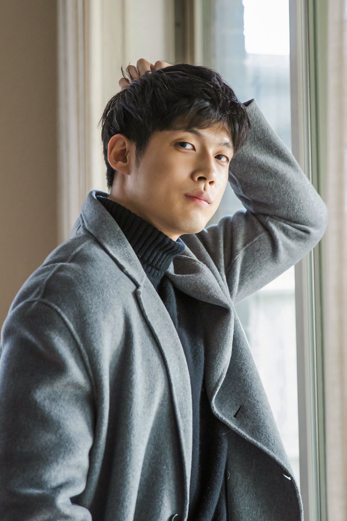 Young Asian man in gray wool coat and turtleneck poses thoughtfully by a window with hand in hair.