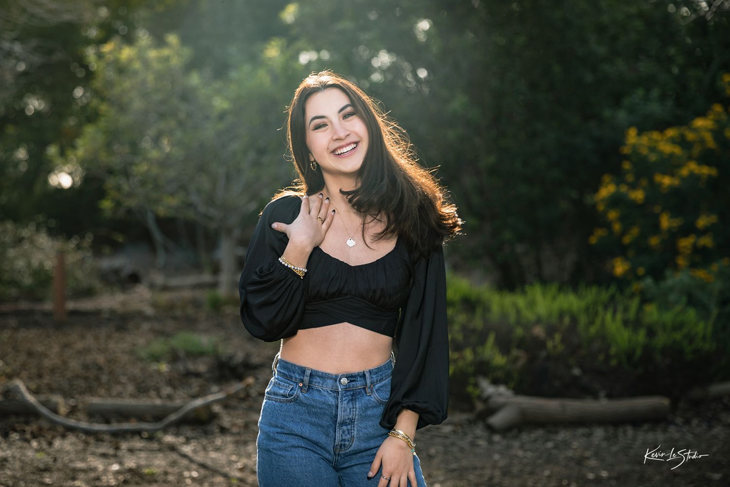 Smiling young woman in black crop top and jeans laughs outdoors in a sunlit forest setting.