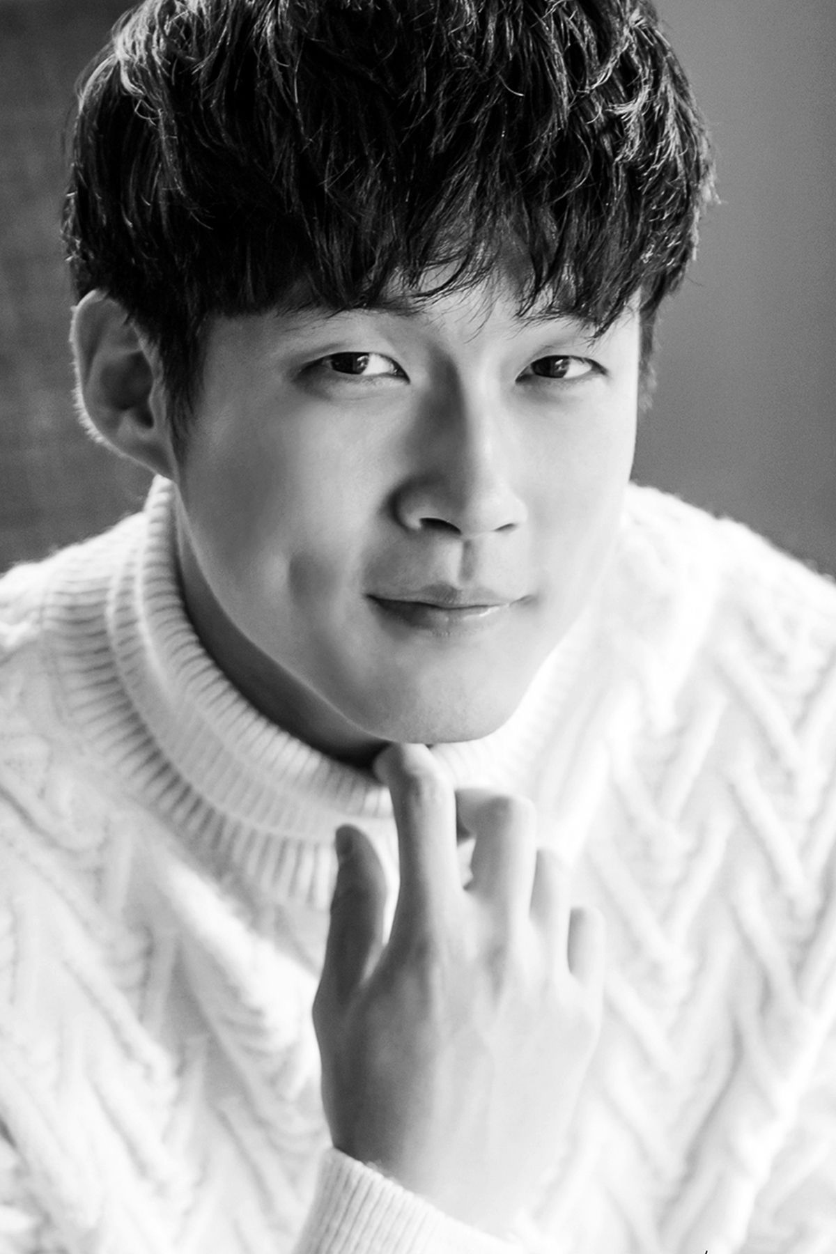 Black and white portrait of a smiling Asian man in white cable-knit sweater with hand near chin.
