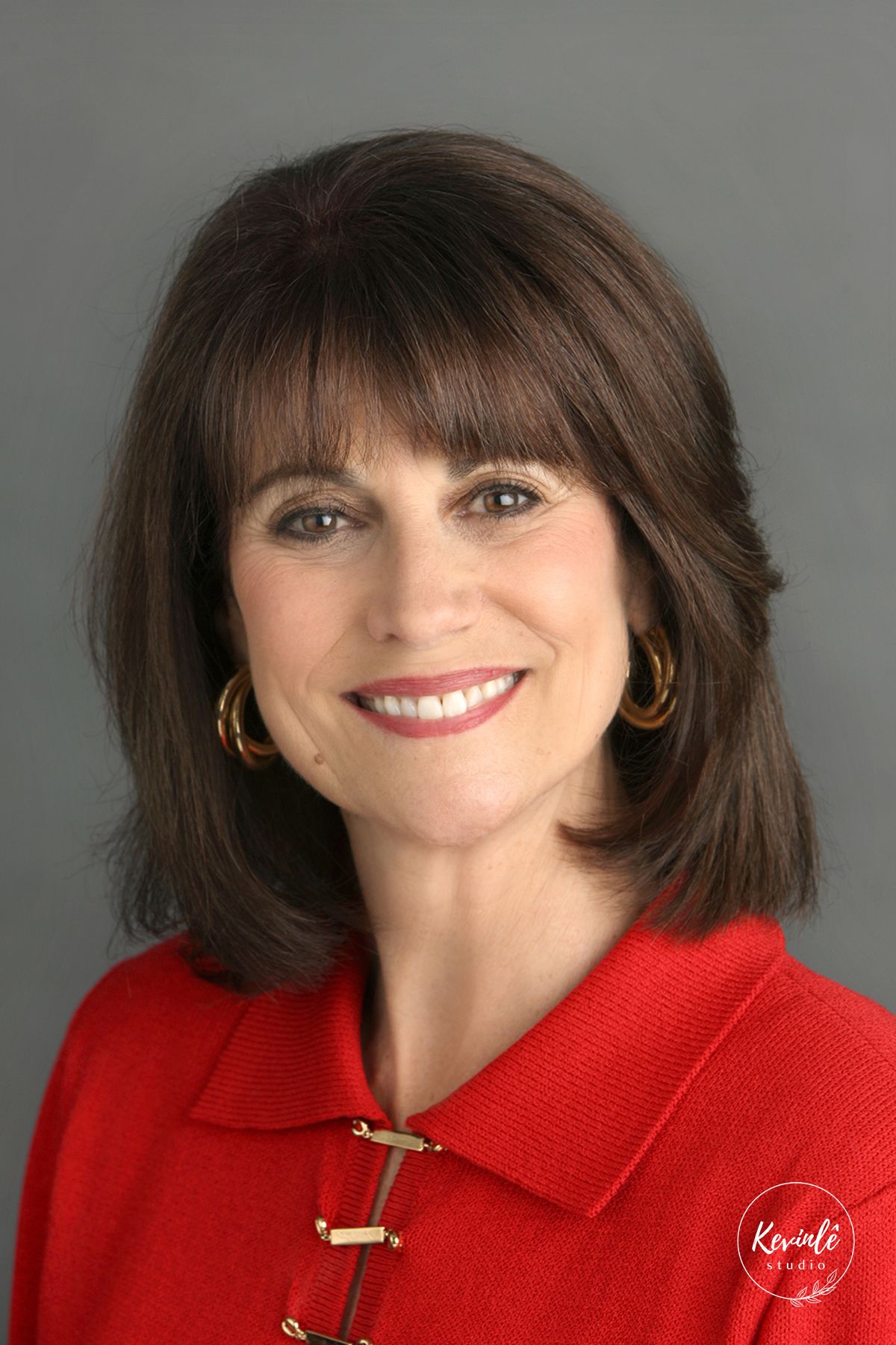 Professional headshot of a smiling brunette woman with bangs wearing a red blazer and gold hoop earrings.
