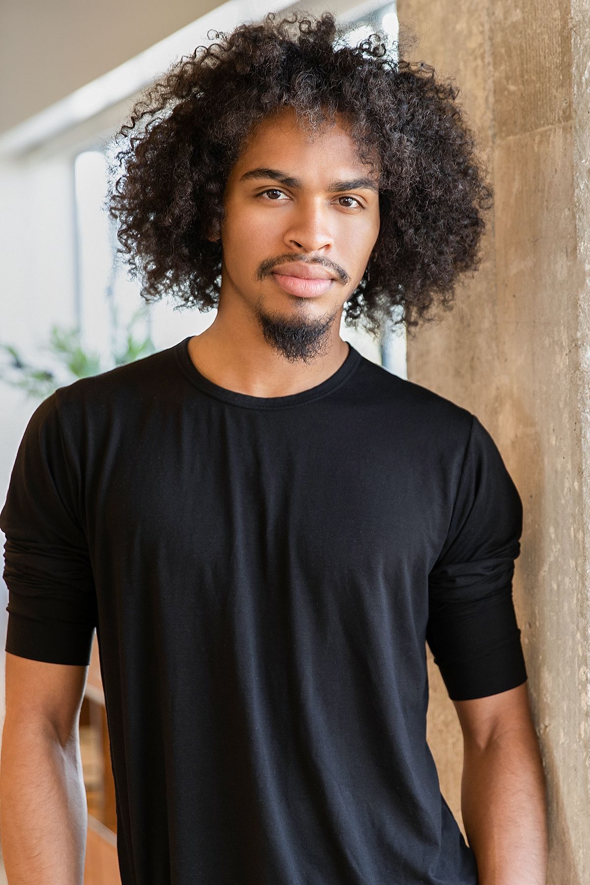 Young Black man with curly afro hair and beard wearing a black long-sleeve t-shirt, leaning against a wall.