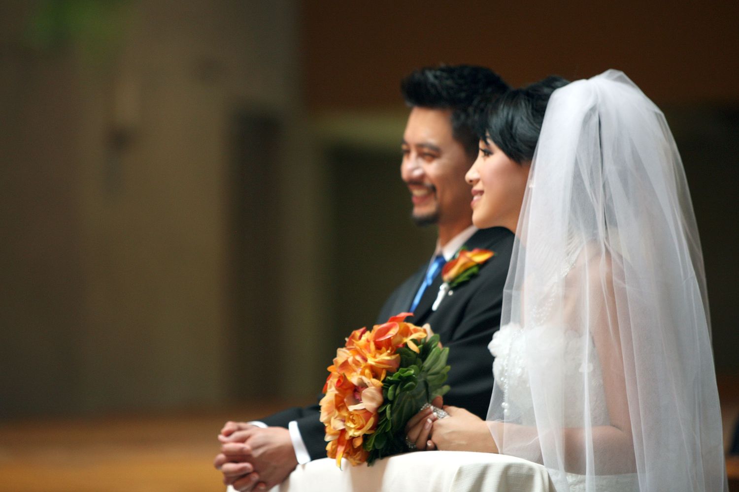 Smiling bride in white veil holding orange bouquet sits beside groom in suit at wedding ceremony.