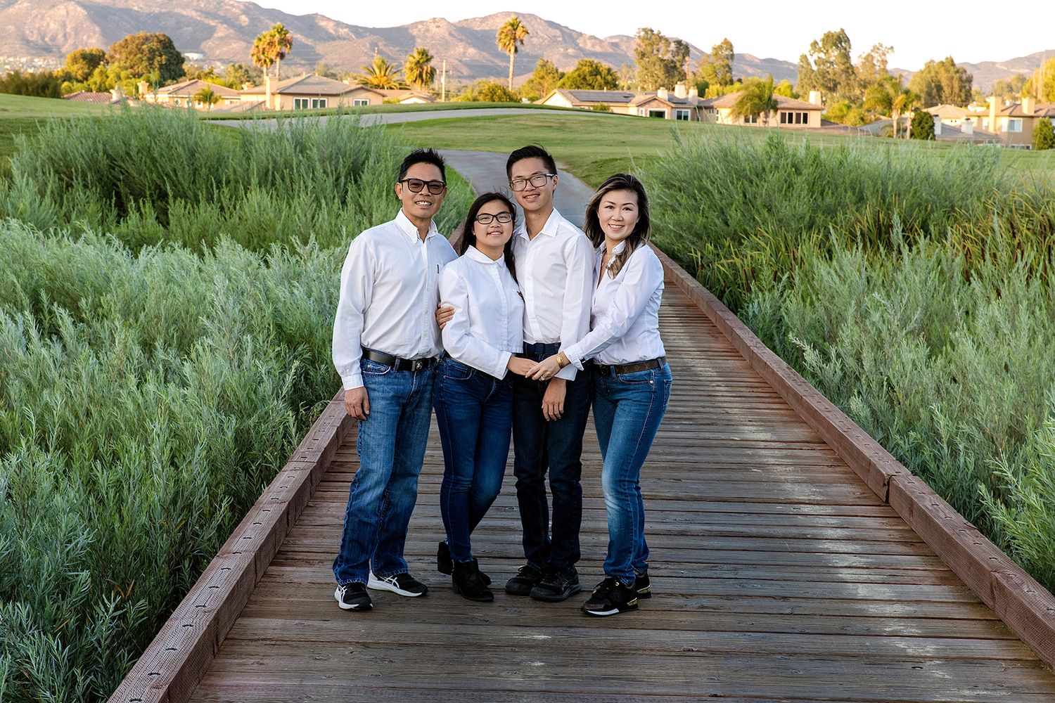 Family of four posing on wooden boardwalk path surrounded by lush greenery with mountains in background at golden hour.