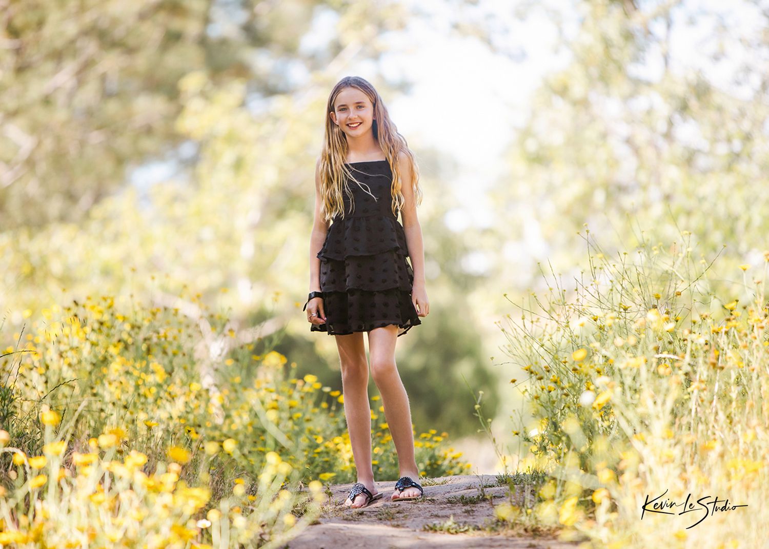 A young girl in a black ruffled dress stands on a rock surrounded by yellow wildflowers in a sunny outdoor setting.