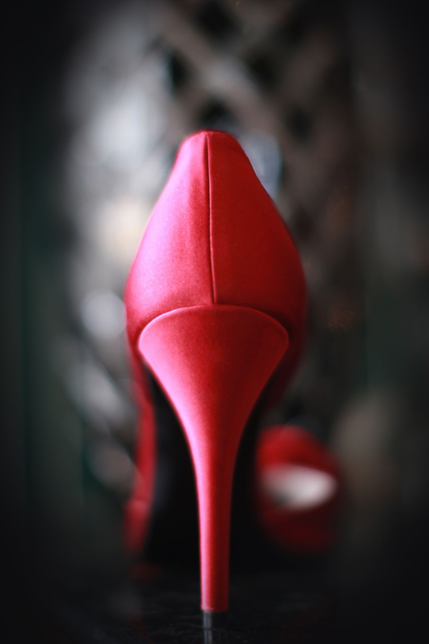 Close-up of a vibrant red satin stiletto high heel shoe photographed from behind on a dark background.
