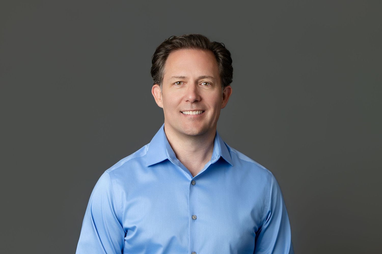 Professional headshot of a smiling man in a light blue dress shirt against a gray background.