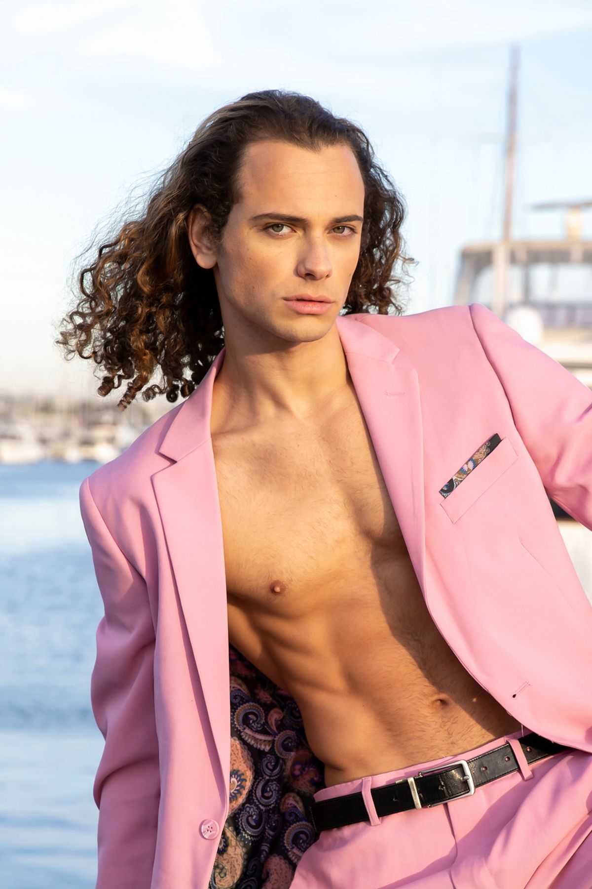 Young man with curly hair wearing an open pink blazer suit with no shirt, posing near a marina.