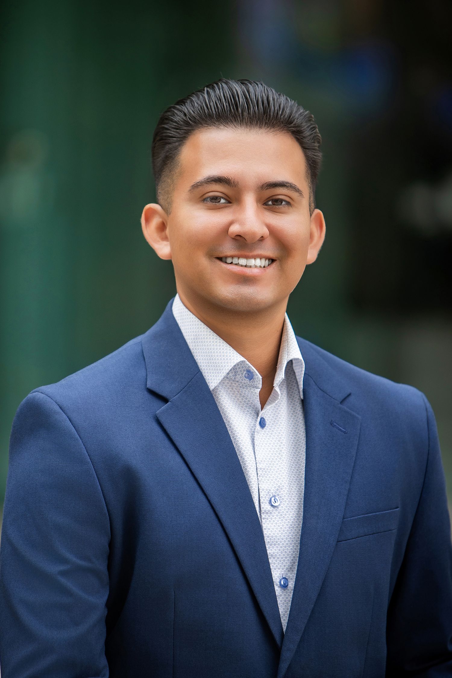 Professional headshot of a smiling young man in a navy blue blazer and white dress shirt outdoors.