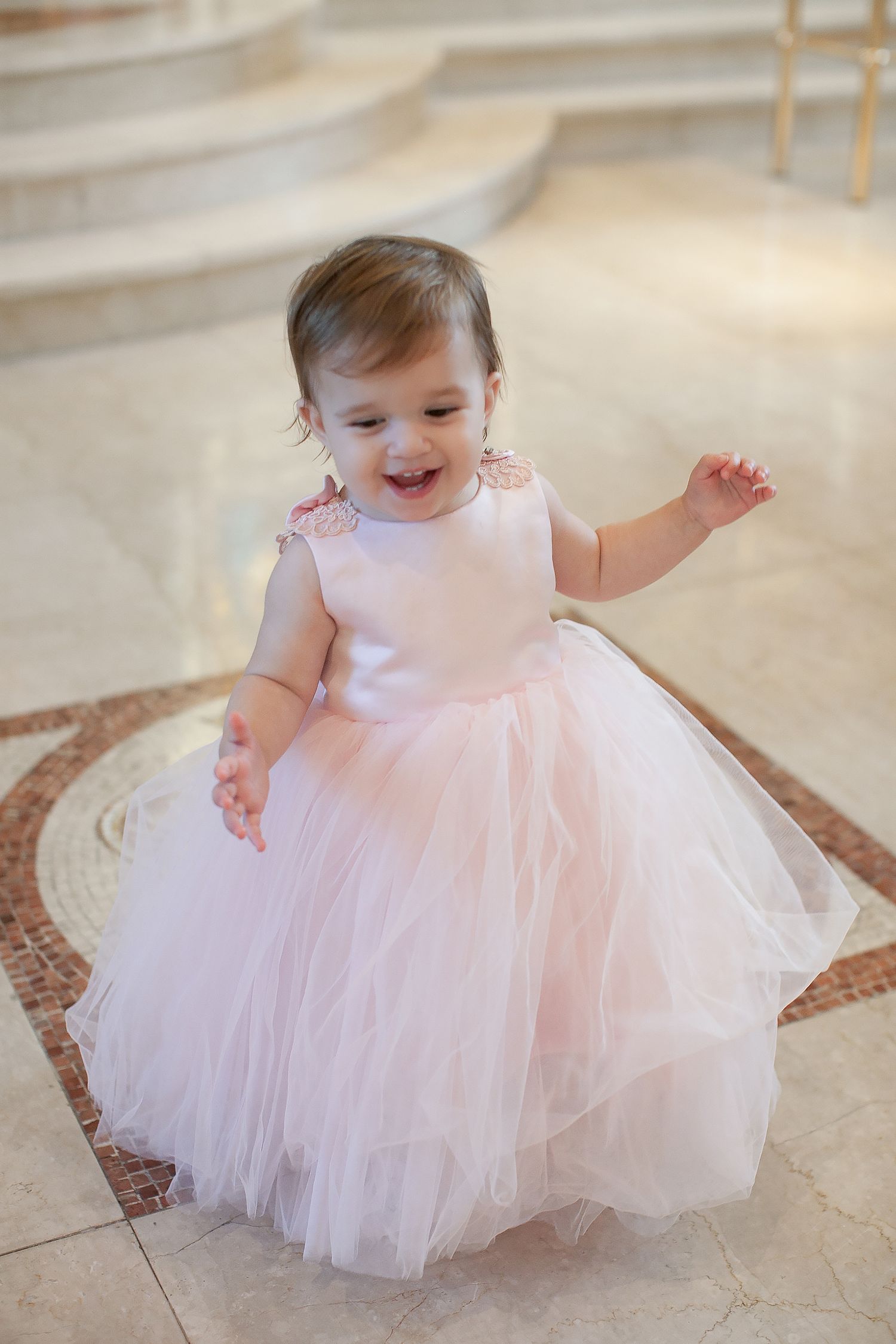 Adorable baby girl laughing joyfully while wearing an elegant pink tulle ball gown dress indoors.