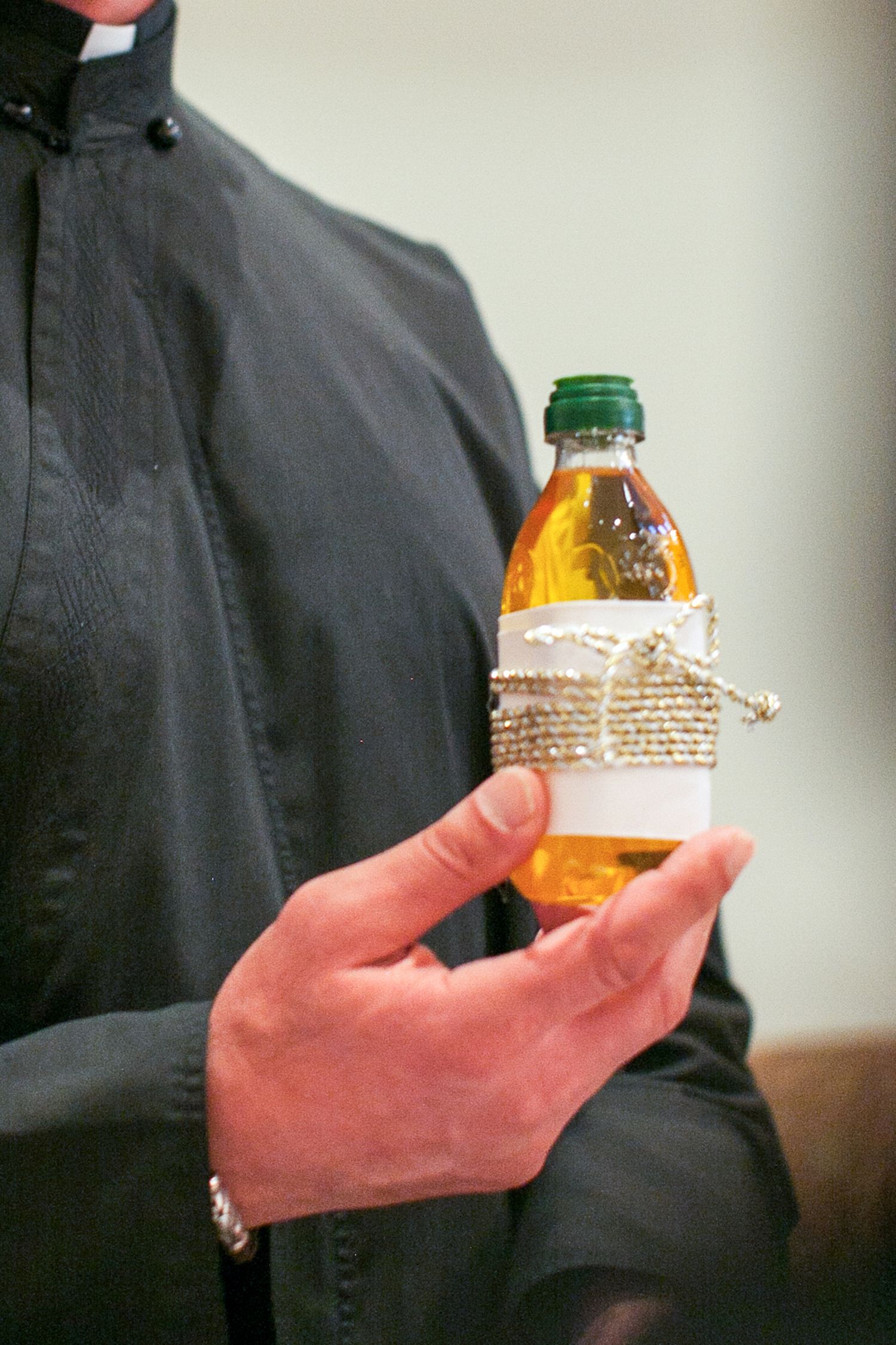 Person in black clergy shirt holding a decorative small bottle of amber liquid with twine and lace wrap.