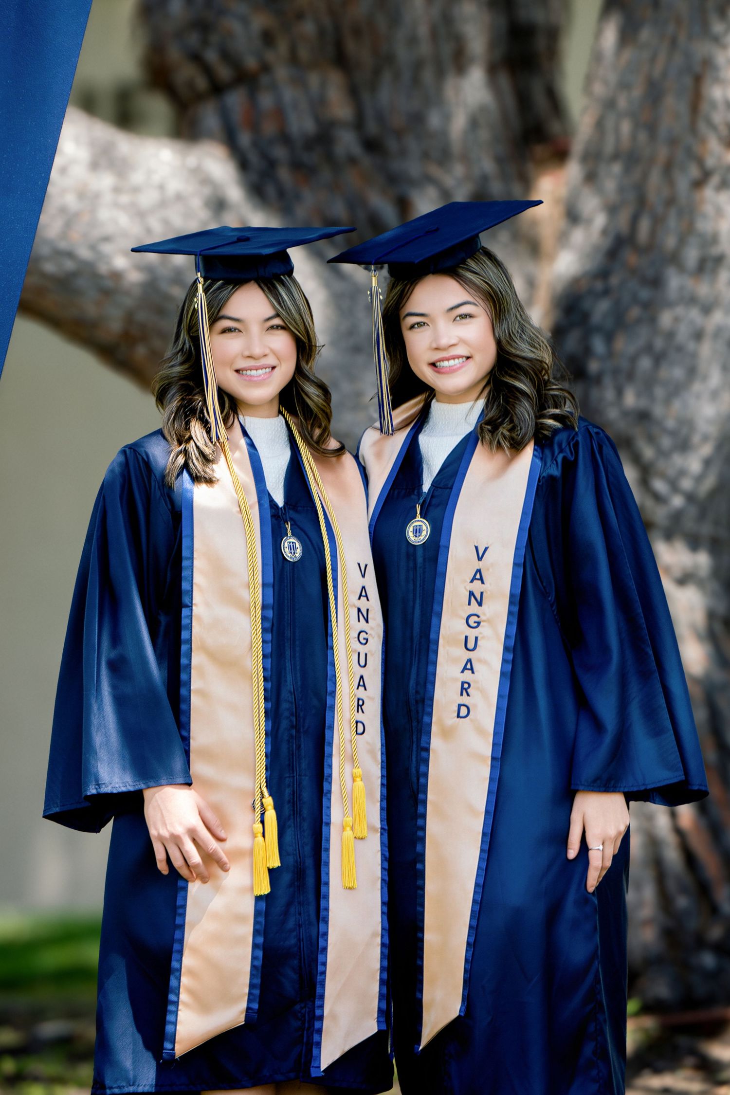 Two smiling women in blue Vanguard graduation gowns and caps pose together outdoors for graduation photos.