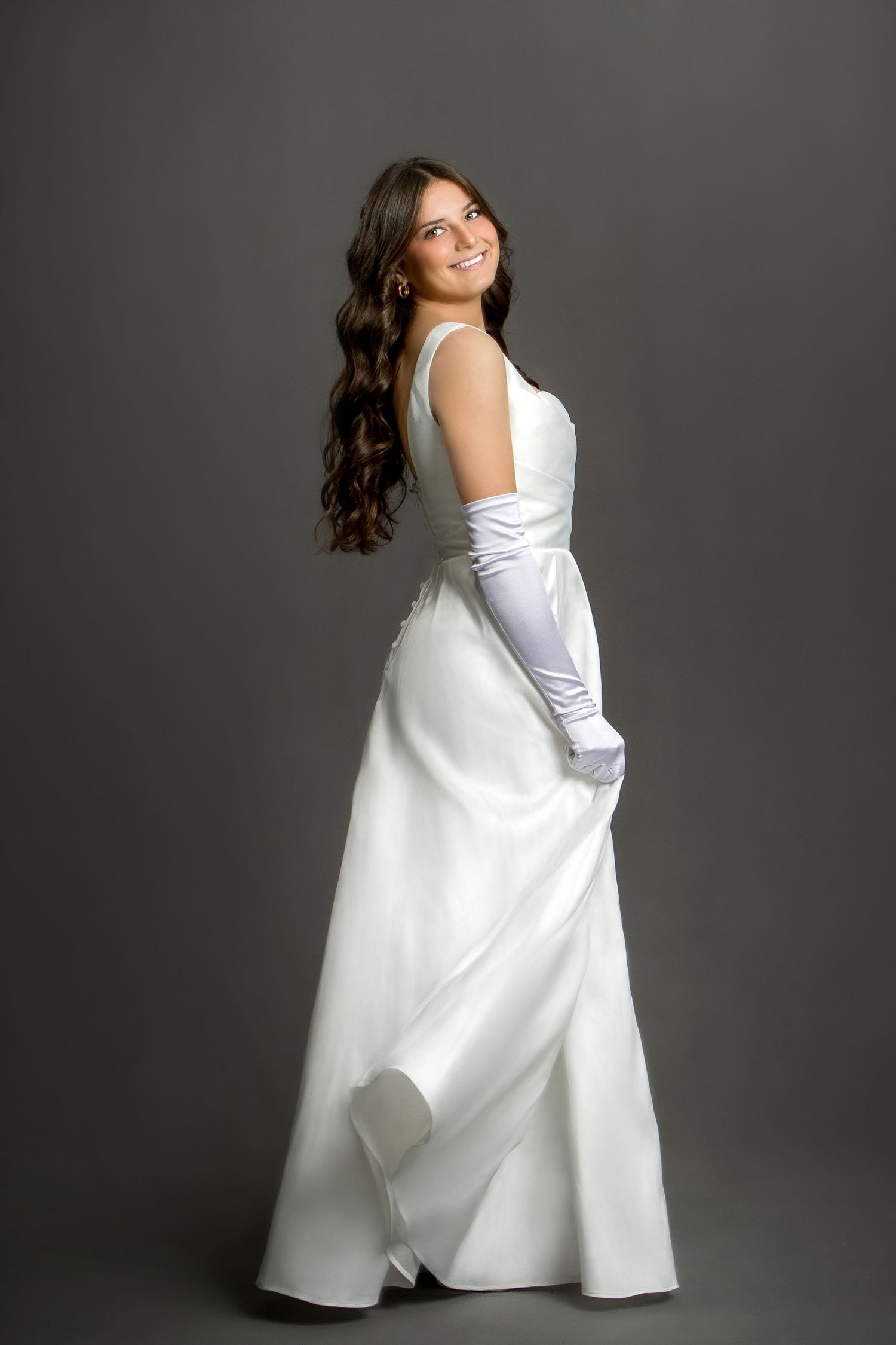 Smiling brunette woman in elegant white satin gown and long white gloves posing gracefully against gray background.