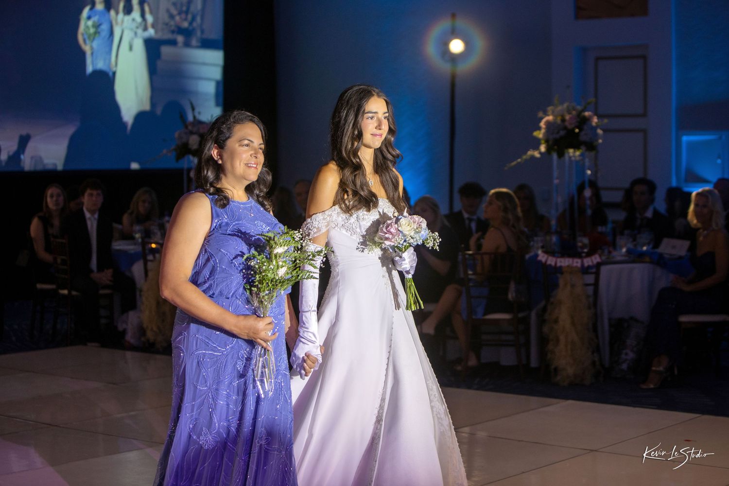 Two elegantly dressed women walk together at a formal event, one in purple and one in a white gown, holding bouquets.