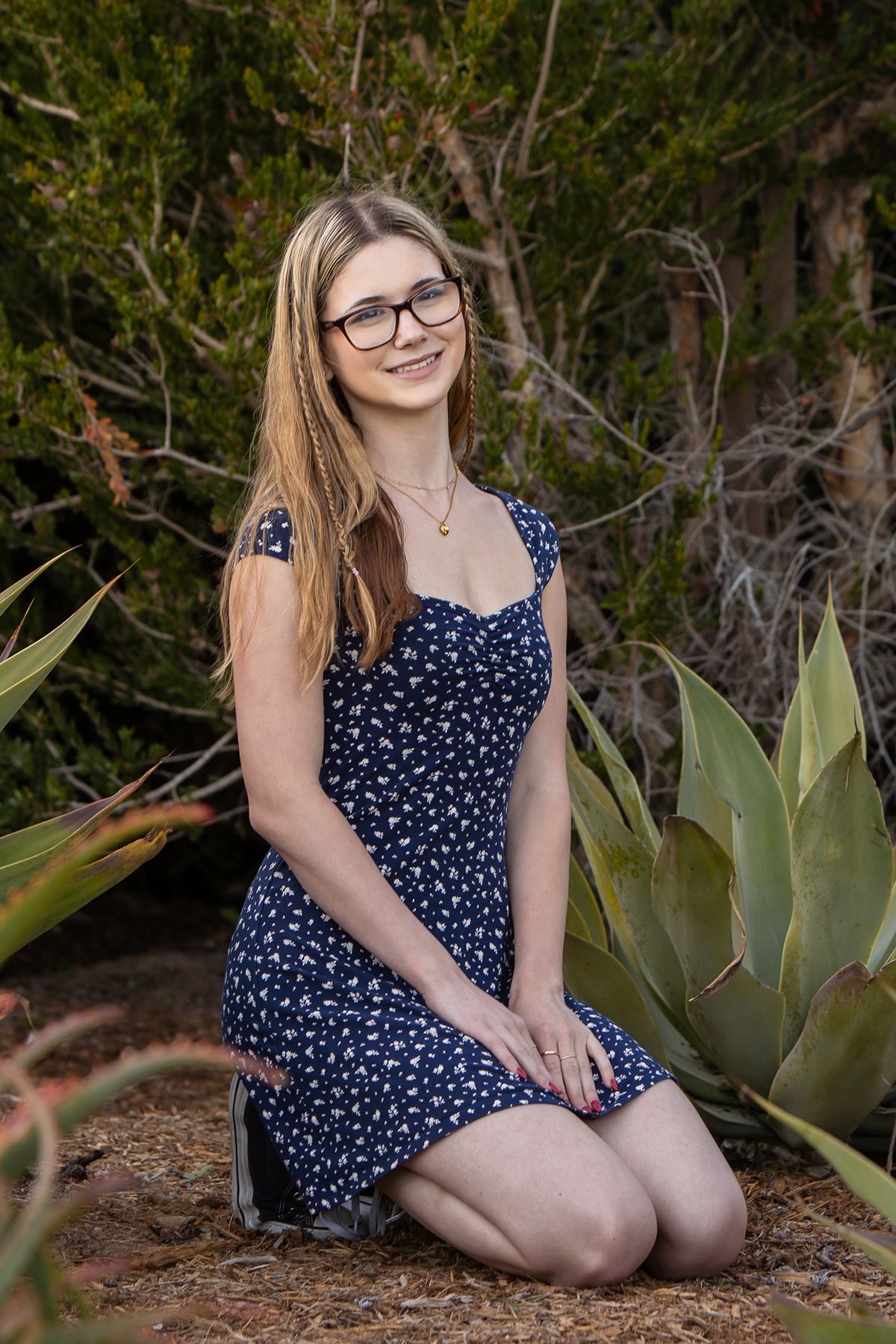 Blonde woman in navy floral dress kneels beside agave plants in a garden landscape.