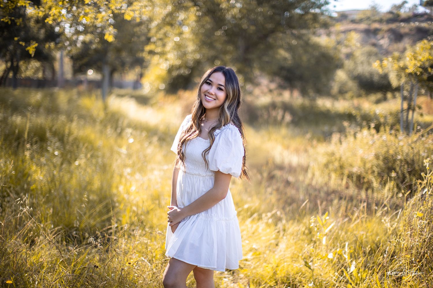 Young brunette woman in white puff sleeve dress smiling in sunlit golden meadow with trees.