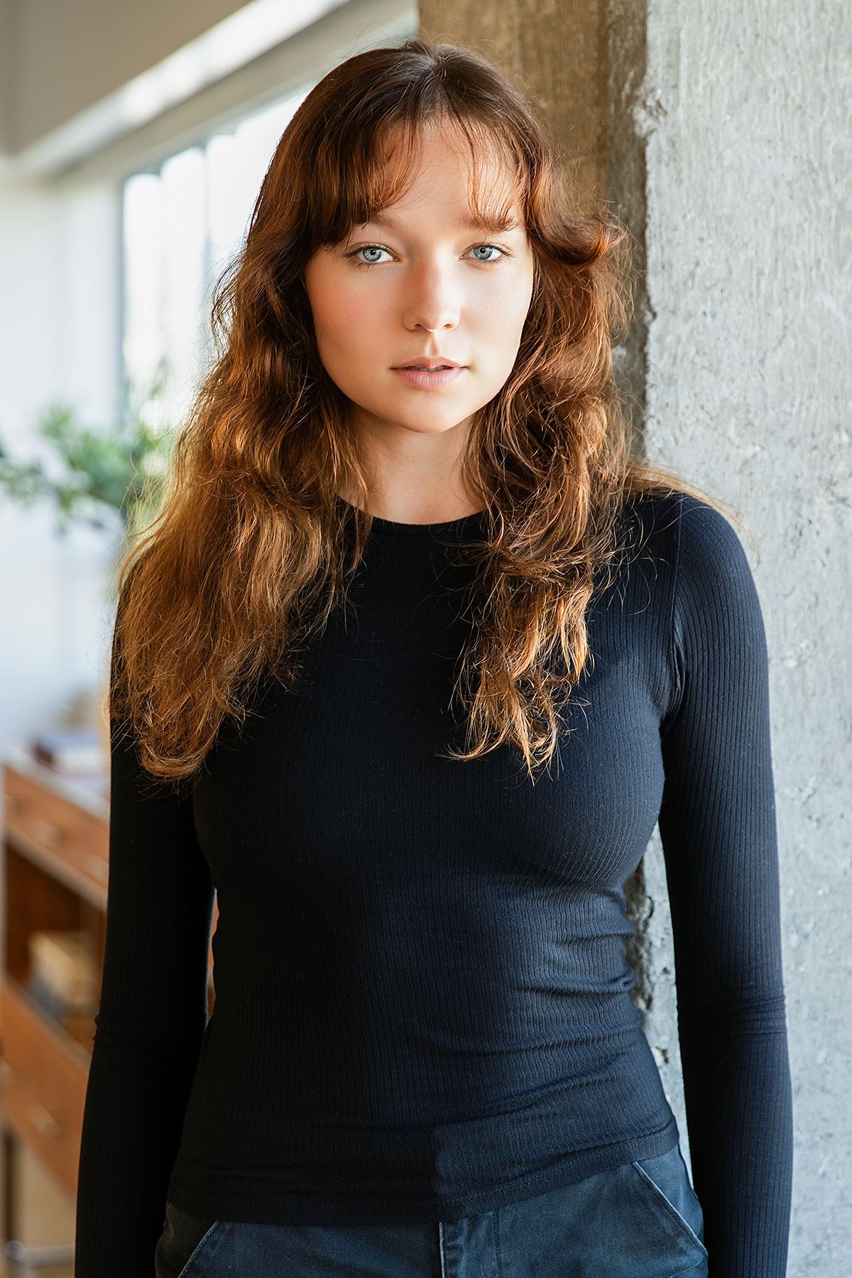 Young woman with wavy auburn hair and bangs wearing a black ribbed long-sleeve top, posing against a concrete wall.